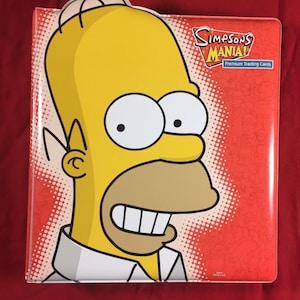 May include: A red binder with a cartoon image of Homer Simpson on the front. The binder is for Simpsons Mania! Premium Trading Cards.