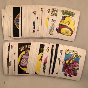 May include: A stack of vintage trading cards featuring various cartoon superheroes. The cards are white with colorful illustrations and text. Some of the characters include "The Incredible Bulk", "Captain Clod", and "Great God".