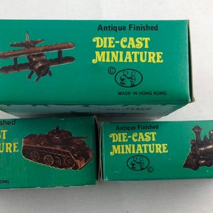May include: Three boxes with green backgrounds and white text. Each box features a different die-cast miniature: a biplane, a tank, and a train. The boxes all have the text "Antique Finished Die-Cast Miniature" and "Made in Hong Kong".