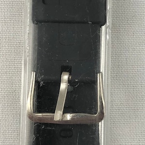 Puede incluir: Una correa de reloj negra con una hebilla plateada en un paquete de pl&aacute;stico transparente. El paquete tiene una etiqueta azul con el texto "SPORTSTRAP TM K-18 FOR CASIO DATABANK 7/8" 22mm 395".