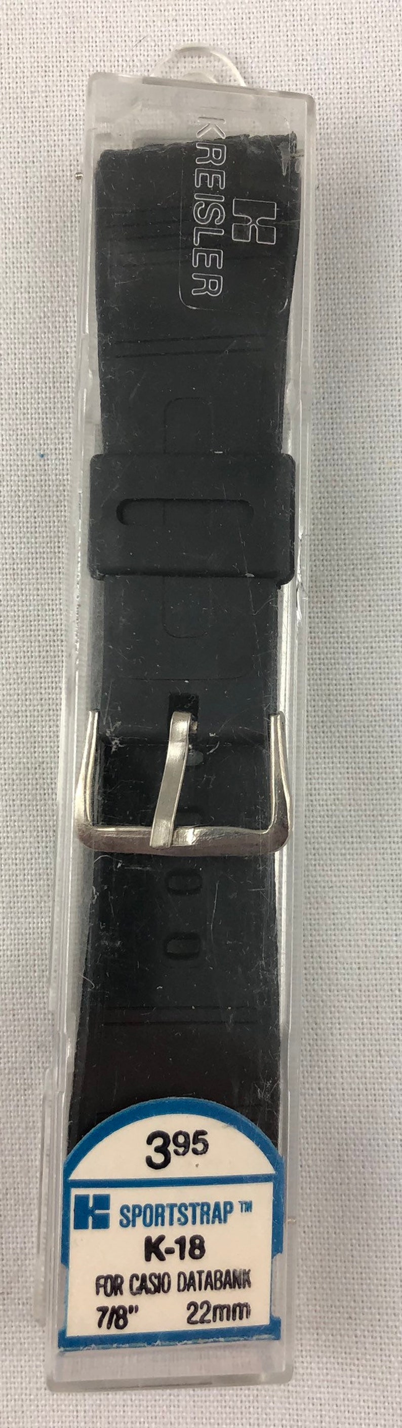 Puede incluir: Una correa de reloj negra con una hebilla plateada en un paquete de pl&aacute;stico transparente. El paquete tiene una etiqueta azul con el texto "SPORTSTRAP TM K-18 FOR CASIO DATABANK 7/8" 22mm 395".