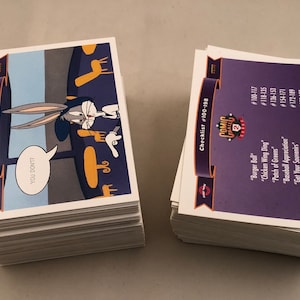 May include: Stacks of trading cards featuring Bugs Bunny from the "Comic Ball 2" series. One stack shows Bugs Bunny with a speech bubble saying "YOU DON'T?". The other stack is a checklist with card numbers and titles.