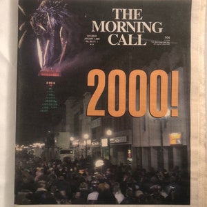 May include: A newspaper front page with the headline "The Morning Call" and the year "2000!" in large, orange text. The image shows a nighttime scene with fireworks in the background and a crowd of people gathered in the street.