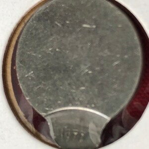May include: Close-up of a silver coin with a smooth, slightly textured surface. The coin has a rounded edge and a slightly raised rim.
