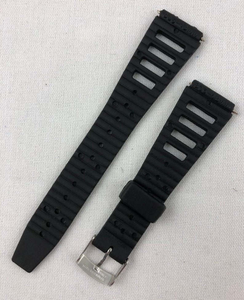 May include: Black rubber watch band with a silver buckle. The band has a textured surface and a series of holes for ventilation.