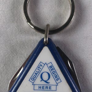 May include: A blue and silver triangle-shaped keychain with a white background. The keychain has a silver ring and the text "Quality Begins Here" with a large "Q" in the center. The text "Bell Atlantic" is below the "Q".