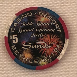 May include: A casino chip with a black background and colorful fireworks design. The chip reads "Casino Resort Table Games Grand Opening 2010 Sands Bethlehem, PA" and has a $5 value.
