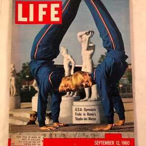 May include: The cover of a vintage "LIFE" magazine from September 12, 1960, features two gymnasts in blue tracksuits performing a handstand. The magazine's title is in a red box. The text reads "U.S.A. Gymnasts Frolic in Rome's Stadio dei Marmi."