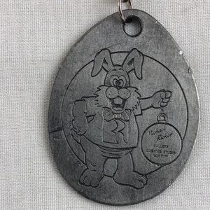 May include: A silver-toned metal keychain featuring a cartoon rabbit holding a Christmas ornament. The rabbit is wearing a blue shirt with a red letter "R" on it. The keychain also includes the text "Michael Ricker Gallery Casting Studio Auburn".