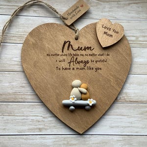 Op de afbeelding: Houten hartvormige plaquette met het woord "Mum" en een gegraveerde liefdesboodschap. Een klein hartje met "Love You Mum" is bevestigd. Voorzien van een steenkunstpaar op een klein skateboard. Inclusief een jute touwhanger.