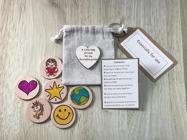 K&ouml;nnte beinhalten: Ein kleiner Jutesack gef&uuml;llt mit Holztoken, jedes mit einem anderen Design. Der Sack hat ein herzf&ouml;rmiges Etikett mit der Aufschrift "Ein kleiner Beutel voller Liebe f&uuml;r meine Enkelin". Eine Karte mit dem Text "Speziell f&uuml;r dich" und einer Liste der Bedeutungen der Token ist ebenfalls enthalten.
