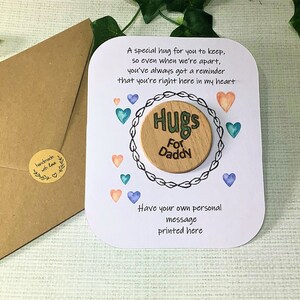Puede incluir: Una tarjeta blanca con un mensaje sobre abrazos para un padre. La tarjeta tiene un c&iacute;rculo de madera con las palabras "Hugs for Daddy" en &eacute;l. La tarjeta est&aacute; decorada con corazones de acuarela en tonos de azul, verde y rosa.