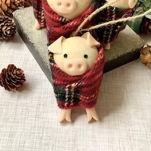 May include: Three small pig ornaments wrapped in red and black tartan fabric. The pigs are made of a light brown material and have small pink snouts.