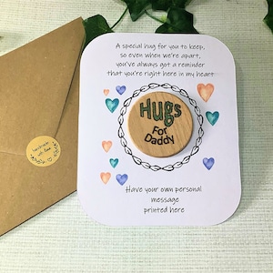 Puede incluir: Una tarjeta blanca con un mensaje sobre abrazos para un padre. La tarjeta tiene un c&iacute;rculo de madera con las palabras "Hugs for Daddy" en &eacute;l. La tarjeta est&aacute; decorada con corazones de acuarela en tonos de azul, verde y rosa.