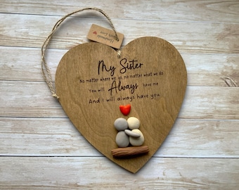 May include: Wooden heart-shaped plaque with the text "My Sister" and a heartfelt message. Features a small red heart and pebble figures. Includes a jute string for hanging.