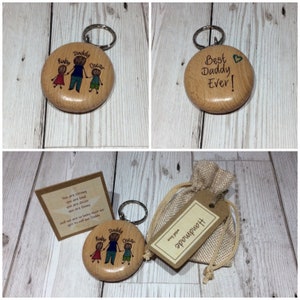May include: A wooden keychain featuring a hand-drawn illustration of a father and two children. The keychain is engraved with the words "Ruby, Daddy, Oscar". The keychain is packaged in a small hessian bag with a tag that reads "Handmade with love".