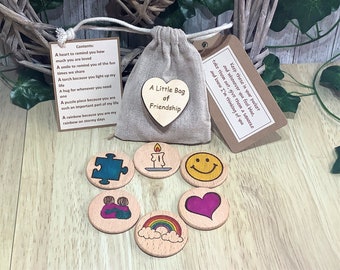 May include: A small burlap bag with a heart-shaped wooden tag that reads "A Little Bag of Friendship". The bag contains six small wooden tokens with various designs, including a puzzle piece, a candle, a smiley face, two people hugging, a heart, and a rainbow.