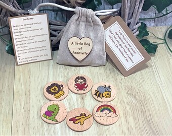 May include: A small burlap bag with a wooden heart that reads "A little Bag of Positivity." The bag is accompanied by wooden tokens with colorful illustrations of a lion, hedgehog, bee, dinosaur, rainbow, and superhero. Text on tags.