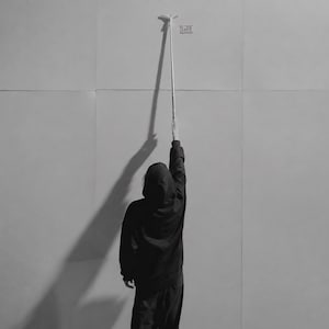 May include: A black and white image showing a person wearing a dark hooded sweatshirt and pants, standing with their back to the camera and reaching up to touch a long, thin, vertical measuring tool against a plain, light-colored wall surface.