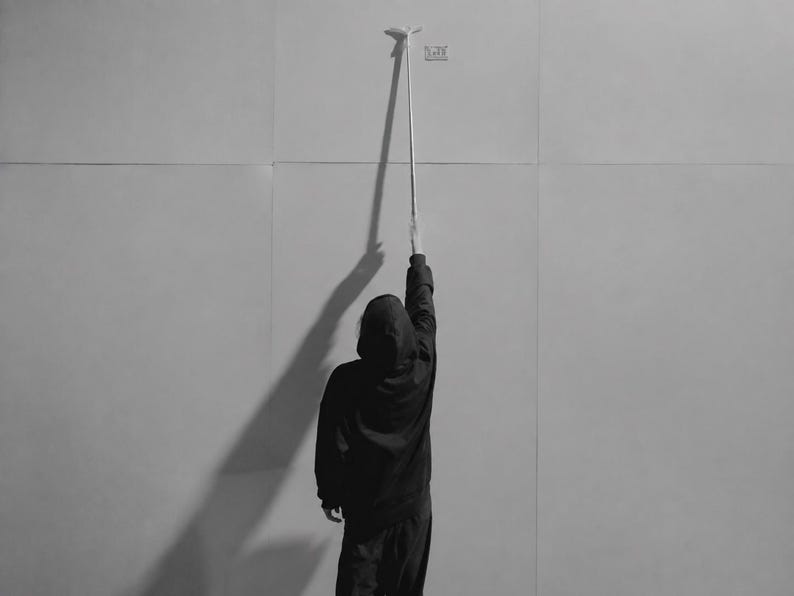 May include: A black and white image showing a person wearing a dark hooded sweatshirt and pants, standing with their back to the camera and reaching up to touch a long, thin, vertical measuring tool against a plain, light-colored wall surface.