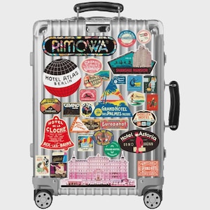 Könnte beinhalten: Ein silberner Rimowa-Koffer mit Vintage-Reiseaufklebern. Die Aufkleber zeigen Hotelnamen und Logos aus verschiedenen Orten, darunter Berlin, Shanghai und Dresden. Der Koffer hat einen Teleskopgriff und Räder.