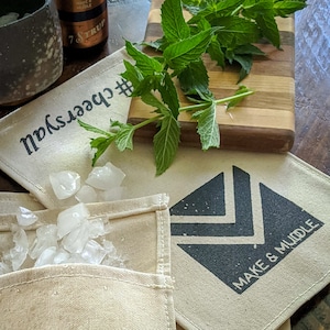 May include: Two white cotton bags with black printed designs. One bag has the text "#CheersYall" and the other has a black triangle design with the text "Make & Muddle". The bags are on a wooden surface with a bunch of fresh green mint leaves and ice cubes.