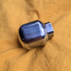 May include: A small, rounded, square-shaped metal object with a polished, reflective silver surface. The item features a smooth, curved body and a distinct, slightly raised cylindrical neck opening at one end, showing a metallic finish.