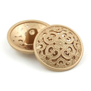 May include: Two gold-toned metal buttons with an intricate, ornate, and lacy design. The buttons are circular and have a raised, textured surface.
