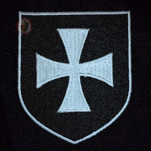 May include: Black embroidered patch with a white Maltese cross design inside a shield shape. The patch has a white border and is on a black background. Suitable for jackets, bags, or other clothing items.