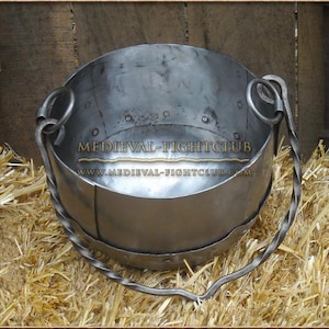 May include: A metal pot with a twisted handle. The pot is made of dark metal and has a rounded bottom. The handle is made of twisted metal and is attached to the pot with two metal rings. The pot is sitting on a bed of straw.
