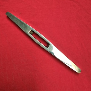 Könnte beinhalten: Eine silberfarbene Metallklinge mit einem rechteckigen Loch in der Mitte. Die Klinge ist an einem Ende spitz und scharf und am anderen Ende abgerundet.