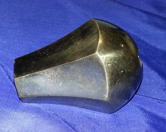 May include: A metallic, geometric object with a polished, reflective surface. The object has a multifaceted design with a rounded end and a flat base. The colour is a dark, silvery grey, and it sits on a blue fabric.