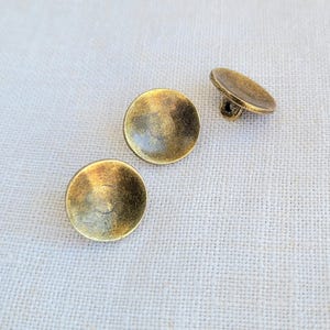 Op de afbeelding: Drie ronde, goudkleurige knopen met een licht verhoogd, gewelfd midden. De knopen hebben een glad, licht getextureerd oppervlak.