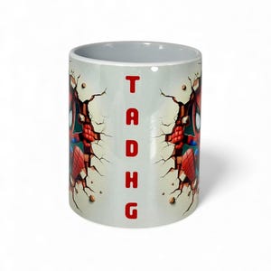 May include: White ceramic mug featuring a Spider-Man design. The design shows Spider-Man breaking through a wall, with the letters "TADHG" in red. The mug has a gray interior.