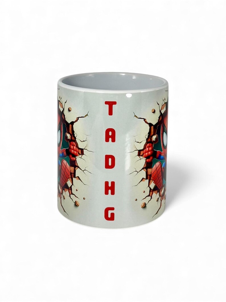 May include: White ceramic mug featuring a Spider-Man design. The design shows Spider-Man breaking through a wall, with the letters "TADHG" in red. The mug has a gray interior.