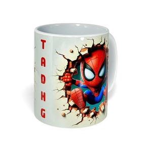 May include: White ceramic mug featuring a cartoon Spider-Man character breaking through a wall. The character is in a red and blue suit. The word "TADHG" is printed vertically in red on the side of the mug.