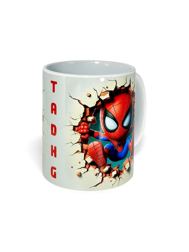 May include: White ceramic mug featuring a cartoon Spider-Man character breaking through a wall. The character is in a red and blue suit. The word "TADHG" is printed vertically in red on the side of the mug.