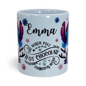May include: White ceramic mug with the name "Emma" in black script. The mug features a design with snowflakes, reindeer, and the text "The North Pole Hot Chocolate Christmas Blend".