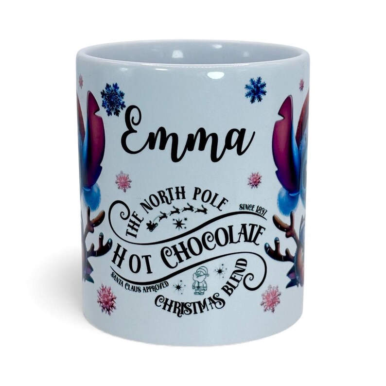 May include: White ceramic mug with the name "Emma" in black script. The mug features a design with snowflakes, reindeer, and the text "The North Pole Hot Chocolate Christmas Blend".