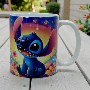 May include: White ceramic mug featuring a colorful illustration of the character Stitch from Lilo & Stitch. The design includes Stitch with a smiling expression, surrounded by flowers and a rainbow background. The mug has a white handle.