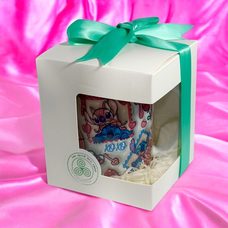 May include: A white gift box with a teal ribbon and bow, showcasing a mug with a cartoon character design. The mug features pink and blue characters, hearts, and the text "xoxo". The box has a window and is filled with shredded paper.