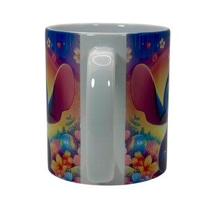 May include: White ceramic mug featuring a colorful cartoon character design. The mug has a white handle and a vibrant wrap-around design with flowers, hearts, and a character's face. The background transitions from blue to yellow.