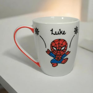May include: White ceramic mug with a red rim and a cartoon image of Spiderman in a red and blue suit. The mug has the name "Luke" written on it in black.