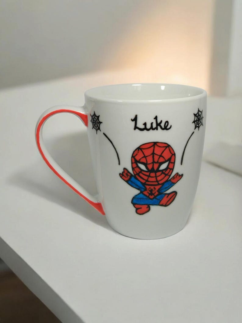 May include: White ceramic mug with a red rim and a cartoon image of Spiderman in a red and blue suit. The mug has the name "Luke" written on it in black.
