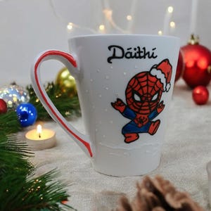 May include: White ceramic mug with a red handle and a red and blue cartoon image of Spiderman wearing a Santa hat. The mug has the name "D&aacute;ith&iacute;" written on it.