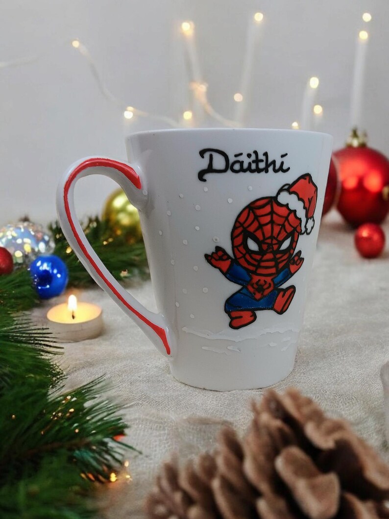 May include: White ceramic mug with a red handle and a red and blue cartoon image of Spiderman wearing a Santa hat. The mug has the name "D&aacute;ith&iacute;" written on it.