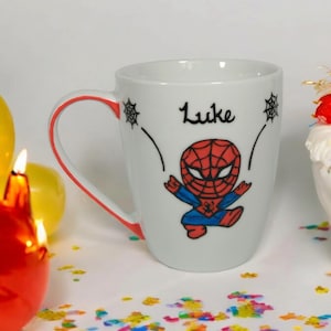 May include: White ceramic mug with a red rim and a cartoon image of Spiderman in red and blue. The mug has the name "Luke" written in black cursive.