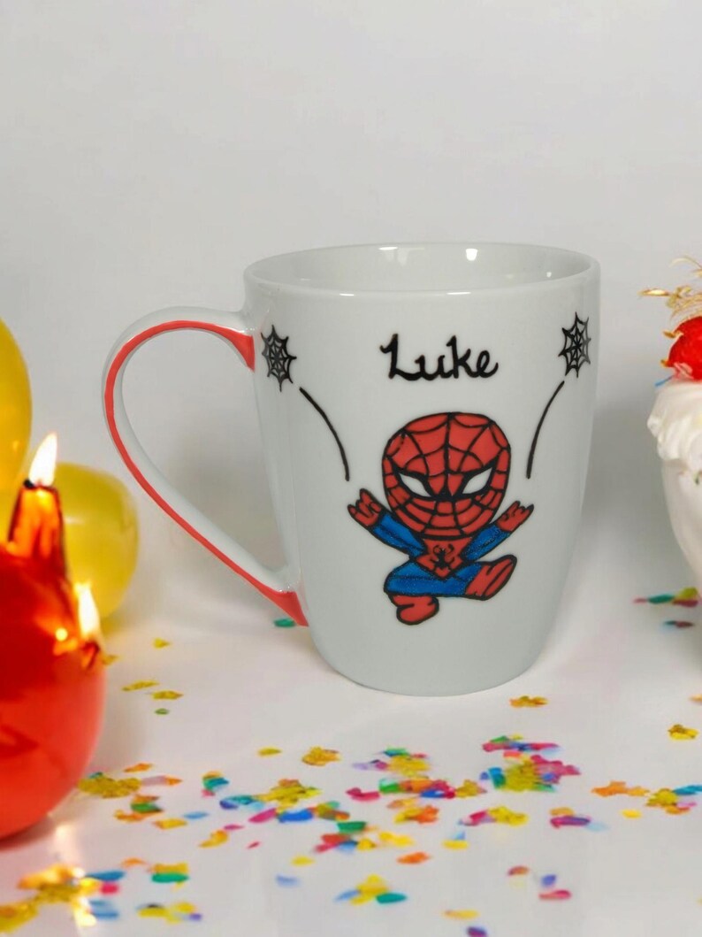 May include: White ceramic mug with a red rim and a cartoon image of Spiderman in red and blue. The mug has the name "Luke" written in black cursive.