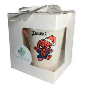 May include: White ceramic mug with a hand-painted design of Spiderman wearing a Santa hat. The mug is in a white gift box with a white ribbon. The mug is personalised with the name "D&aacute;ith&iacute;".