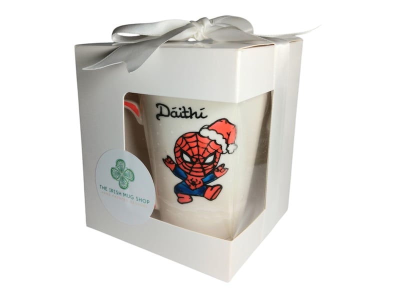 May include: White ceramic mug with a hand-painted design of Spiderman wearing a Santa hat. The mug is in a white gift box with a white ribbon. The mug is personalised with the name "D&aacute;ith&iacute;".